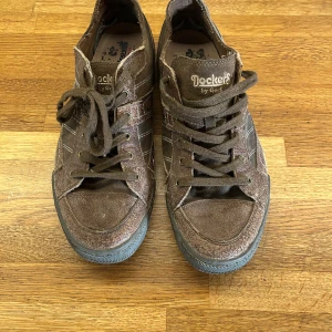 Bruna sneakers från Dockers i mocka och läder - Snygga bruna sneakers från Dockers by Gerli i mocka med vita och bruna detaljer på sidorna. Skorna har snörning, låg profil och en platt sula med mönstrad undersida. Perfekta för en avslappnad streetstil. Sparsamt använda
