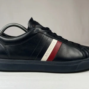 Moncler svarta sneakers i skinn - Snygga svarta sneakers från Moncler i äkta skinn med klassisk låg profil. Skorna har svarta snören och en ikonisk Moncler-logga på plösen. Sidan pryds av en röd och vit rand med Moncler-tryck som ger en sportig och exklusiv känsla. Pris pga ingen original box eller dustbag 