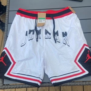 Jordan vita basketshorts med röd detalj - Pris kan diskuteras. Snygga vita Jordan basketshorts med svart och röd rand längs midjan och benen. Stora JORDAN-bokstäver framtill och klassiska Jumpman-loggor på sidorna. Meshmaterial för extra andningsförmåga och svart snörning i midjan. Perfekta för sport eller streetwear.