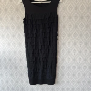 Svart kortklänning från Bruuns Bazaar - Snygg svart kortklänning från Bruuns Bazaar med ärmlös design och rund halsringning. Framsidan har flera lager av volangliknande rynkade band som ger en cool struktur. Perfekt för dig som gillar stilrena och trendiga plagg. Använd fåtal gånger. Rökfritt hem!
