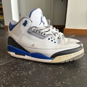 Nike Air Jordan 3 Retro Vit/Blå - Nike Air Jordan 3 Retro sneakers i vitt skinn med blå och grå detaljer. Klassisk siluett med snörning, perforerade partier och ikoniskt elefantmönster på tån och hälen. Jumpman-logga i blått på plösen och blå inslag på sulan. Perfekta för dig som gillar streetwear. Box finns kvar.