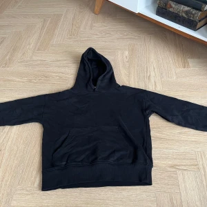 Svart hoodie med ficka, stl 146/152 - En svart hoodie i klassisk modell med stor känguruficka framtill och huva. Tröjan har långa ärmar och ribbade muddar vid ärmslut och nederkant. Perfekt för chill dagar eller när du vill ha en enkel och stilren look. Liten fläck som går bort i tvätten på armen.