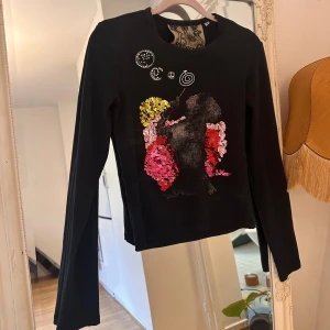 Svart långärmad tröja med paljetter - Svart långärmad tröja med coolt tryck framtill föreställande en person med paljetter i gult, rosa och rött.