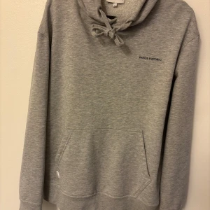 Grå hoodie från Panos Emporio - Snygg grå hoodie från Panos Emporio med diskret logga på bröstet. Tröjan har huva med snörning, känguruficka och långa ärmar. Perfekt för chill dagar eller när du vill ha en enkel och stilren look.