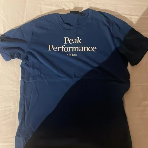 Blå Peak Performance t-shirt - Snygg blå t-shirt från Peak Performance med vit logga och text 'Est. 1986' på bröstet. Klassisk rund hals och korta ärmar, perfekt för en avslappnad och sportig stil. Skön passform och stilren design som funkar året runt.