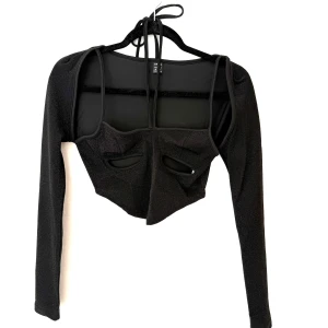 Svart glittrig croppad topp  - Svart glittrig croppad topp med långa ärmar och coola cut-outs framtill. Toppen har knyt i nacken för extra detalj.