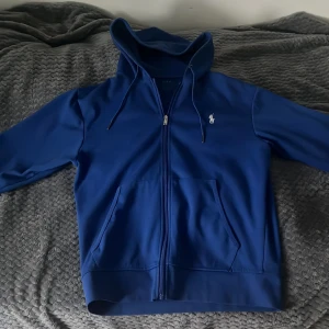 Blå hoodie från Polo Ralph Lauren - Snygg blå hoodie från Polo Ralph Lauren med dragkedja och klassisk logga på bröstet. Tröjan har huva med snörning, två stora fickor fram och långa ärmar. Perfekt för en chill och stilren look.