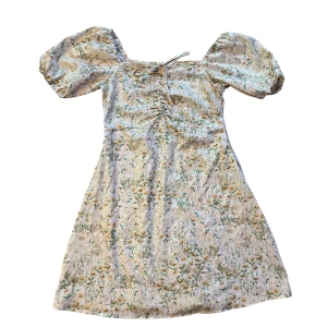 Blommig kortklänning med puffärm - Supersöt kortklänning med blommigt mönster i ljusa toner. Klänningen har puffiga kortärmar, snörning framtill och en lätt A-linjeformad siluett. Perfekt för varma dagar och somriga vibes.