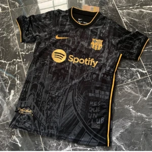 Svart FC Barcelona Nike fotbollströja - FC Barcelona matchtröja i svart med guldiga detaljer och loggor. Tröjan har klubbmärke, Nike Swoosh och sponsortryck i guld, samt guldkantad V-ringning och ärmslut. perfekt för fotbollsträning eller att bära på läktaren.