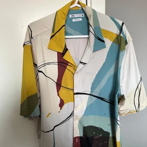 Färgglad kortärmad skjorta från Zara - Säljer en färgglad kortärmad skjorta från Zara med abstrakt mönster i blått, gult, rött och beige. Skjortan har klassisk krage, knäppning framtill och loose fit. Perfekt för dig som gillar unika plagg med mycket färg och stil.