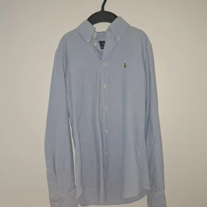 Ljusblå skjorta från Ralph Lauren - Klassisk ljusblå skjorta från Ralph Lauren med button-down krage och broderad logga på bröstet. Skjortan har långa ärmar, knäppning framtill och är tillverkad i mjuk bomull. Perfekt för en clean och stilren look nu till sommaren