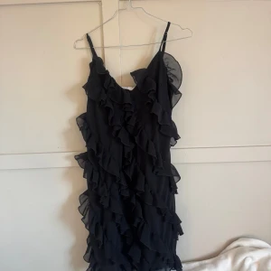 Svart volangklänning från NA-KD - Supersnygg svart kortklänning från NA-KD med tunna axelband och massor av volanger som ger en riktigt cool och lekfull look. Perfekt för dig som vill sticka ut med en feminin och trendig stil.