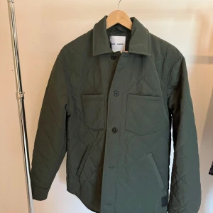 Quiltad grön jacka från Samsøe Samsøe - Snygg quiltad jacka från Samsøe Samsøe i mörkgrönt. Jackan har raka linjer, två bröstfickor, knäppning med knappar och klassisk krage. Perfekt för lager-på-lager och chill höst- eller vårstil.