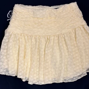 Vit volangkjol från Gina Tricot XS - Supersöt kort kjol från Gina Tricot i vitt med volanger och diskret spetsmönster. Kjolen har en smockad midja med snörning i sidan och är tillverkad i ett lätt och luftigt tyg, perfekt för varma dagar.