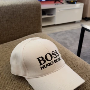 Vit keps från Hugo Boss - Snygg vit keps från Hugo Boss med broderad svart logga framtill. Klassisk böjd skärm och justerbart spänne bak i metall. Perfekt accessoar för en clean och stilren look. Tillverkad i bomull för skön känsla.