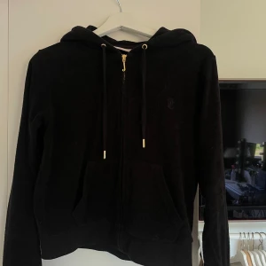 Svart hoodie från Juicy Couture - Svart hoodie från Juicy Couture med dragkedja, huva och snörning. Tröjan har känguruficka framtill och är tillverkad i en mjuk mix av bomull och polyester. Perfekt för en chill och sportig look. Dragkedjan är guldig
