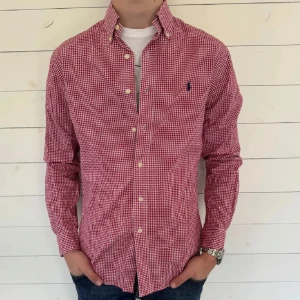 Röd rutig skjorta från Ralph Lauren - Snygg röd och vit rutig skjorta från Ralph Lauren med klassisk button-down krage och broderad logga på bröstet. Skjortan har långa ärmar och knäppning framtill. Perfekt för en stilren och avslappnad look.