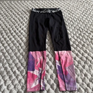 Björn Borg leggings med färgglada detaljer - Snygga leggings från Björn Borg i svart med bred resår i midjan och logga. Nedre delen av benen har ett coolt grafiskt mönster i rosa, lila och vitt. Perfekta för träning eller chill. Stretchigt syntetmaterial som sitter tight och bekvämt.