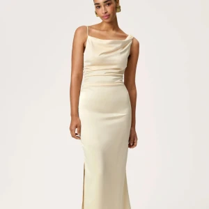 Vanilj gul  långklänning i satin - Elegant långklänning i champagnefärgad satin med smala axelband och en snygg oneshoulder-design. Klänningen har en slits på sidan och figurnära passform som ger en lyxig och stilren look. Perfekt för speciella tillfällen. Den är slutsåld på deras hemsida.