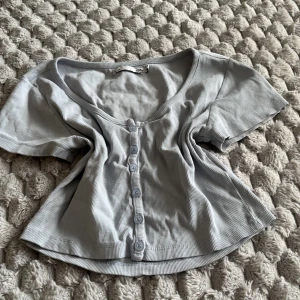 Ljusgrå ribbad topp från 157 - Supersöt ljusgrå ribbad topp från 157 med korta ärmar och knappar hela vägen framtill. Toppen har en snygg v-ringning och croppad passform som ger en trendig vibe. Perfekt att matcha med jeans eller kjol för en chill look.