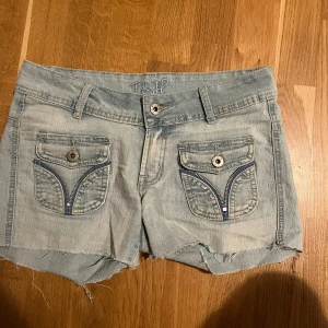 Ljusblå jeansshorts med dragkedjedetaljer - Ljusblå jeansshorts från T&H med råa kanter och snygga dragkedjedetaljer på fickorna. Shortsen har låg midja, bälteshällor och knäppning fram. Materialet är en mix av polyester och elastan för extra stretch. Perfekta för varma dagar och en avslappnad look.