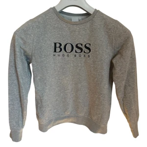 Grå sweatshirt från Hugo Boss - Snygg grå sweatshirt från Hugo Boss med svart logga framtill. 138 cm - 10 år jättefint skick. Klassisk rund halsringning och långa ärmar. Mjuk och skön bomullsblandning som passar perfekt till jeans eller joggers. Enkel men stilren design för en clean look.