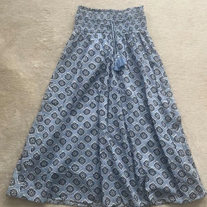 Blå långkjol med mönster från Gina Tricot - Supersöt långkjol från Gina Tricot i blått och vitt med snyggt mönster. Kjolen har smock i midjan, knytband med tofsar och är gjord i ett lätt bomullstyg. Perfekt för en chill sommardag eller när du vill ha en boho vibe.