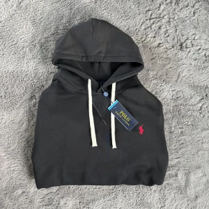 Svart hoodie från Polo Ralph Lauren - Svart hoodie från Polo Ralph Lauren med vit snörning och klassisk röd broderad logga på bröstet. Tröjan har huva och är tillverkad i mjuk bomull, perfekt för en chill och stilren look.