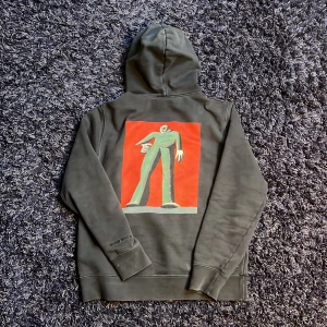 Svart hoodie Wood Wood x T.R. Marshall - Säljer en svart hoodie från Wood Wood i samarbete med T.R. Marshall. Tröjan har en stor färgglad print på ryggen, broderad logga på bröstet och ärmen samt klassisk känguruficka och huva. Perfekt för dig som gillar streetwear och unika prints. Hör av dig vid minsta fundering🙌