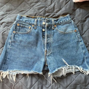 Levi's 501 jeansshorts med fransig kant - Klassiska blå jeansshorts från Levi's modell 501 med hög midja och rå, fransig kant. Shortsen har knäppning framtill, fem fickor och den ikoniska läderpatchen bak. Perfekta för sommaren och ger en avslappnad vintagekänsla. Strolek W26 så känns som Storlek small for dam 