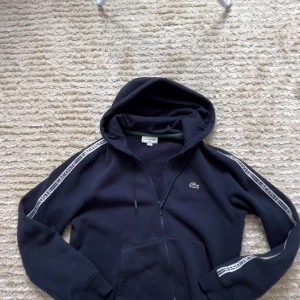 Marinblå hoodie från Lacoste Storlek M - Snygg marinblå hoodie från Lacoste med dragkedja och huva. Klassisk passform, vita logoband längs ärmarna och den ikoniska krokodilloggan på bröstet. Perfekt för en sportig och avslappnad stil.