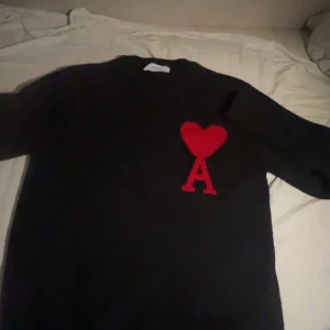 Svart stickad tröja från Ami Paris - Svart stickad tröja från Ami Paris med ikoniskt rött hjärta och bokstaven A broderat på bröstet. Klassisk rund halsringning och långa ärmar. Perfekt för dig som gillar stilrena plagg med en twist.
