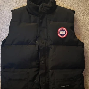 Svart dunväst från Canada Goose - Svart dunväst från Canada Goose med klassisk logga på bröstet. Västen har hög krage, tryckknappar och dragkedja framtill samt två stora fickor. Perfekt för lager-på-lager och riktigt snygg till streetwear eller outdoor-stil.