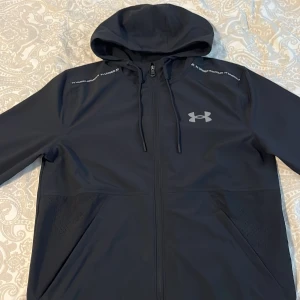 Svart vindjacka från Under Armour - Svart vindjacka från Under Armour med huva och dragkedja hela vägen. Jackan har snörning i huvan, två stora fickor och logga på bröstet. Materialet är lätt och känns sportigt, perfekt för aktiva dagar. Snygga detaljer med text på axlarna.