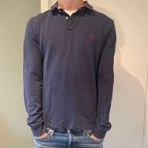Mörkblå långärmad piké från Polo Ralph Lauren - Snygg mörkblå långärmad pikétröja från Polo Ralph Lauren i slim fit. Klassisk krage, knäppning vid halsen och ikonisk röd logga broderad på bröstet. Tillverkad i mjuk bomull som är skön att bära. Perfekt för en clean och stilren look.