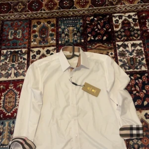 Vit Burberry skjorta med rutiga detaljer - Helt ny snygg vit skjorta från Burberry med klassiska rutiga detaljer på krage, knappslå och ärmslut. Skjortan har broderad logga på bröstet och är långärmad med normal passform. Perfekt för dig som vill ha en stilren look med lyxig känsla.