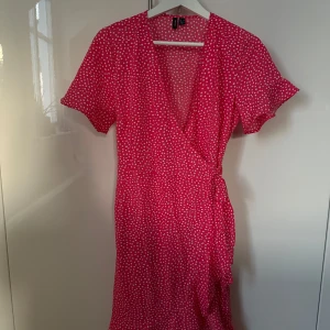 Rosa omlottklänning med vita prickar  - Superfin rosa omlottklänning från Vero Moda med vita prickar. Kort ärm, v-ringning och knyt i midjan för en snygg passform. Perfekt för sommarens alla tillfällen. Lätt och luftig modell som känns både trendig och bekväm.