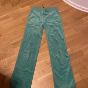 Turkosa mjukisbyxor Juicy Couture - Turkosa mjukisbyxor från Juicy Couture i mjuk sammet med bred resår och snörning i midjan. Byxorna har raka ben, stora fickor fram och bak samt broderad JC-logga på ena fickan. Perfekta för en chill och trendig look.