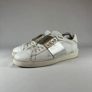 Valentino Open sneakers i vitt och silver - Snygga Valentino Open sneakers i vitt skinn med metallic-silver panel och ikoniska nitar på hälen. Klassisk låg modell med snörning och rund tå. Perfekta för dig som vill ha en clean men ändå lyxig look.