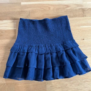 Mörkblå volangkjol med smock - Supersöt mörkblå kortkjol med smockad midja och flera lager volanger. Kjolen har en luftig känsla och passar perfekt till sneakers eller boots. Materialet känns lätt och stretchigt, vilket gör den extra bekväm och snygg.