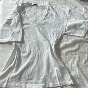 Vit v-ringad t-shirt från Zara - Vit t-shirt från Zara med v-ringning och korta ärmar. Modellen är lite loose och gjord i mjuk bomull, perfekt för en chill och enkel stil. Passar dig som gillar basic men ändå vill ha något snyggt och bekvämt.