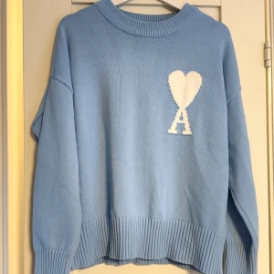 Ljusblå stickad tröja från Ami Paris - Superfin ljusblå stickad tröja från Ami Paris med oversized passform. Tröjan har ett vitt hjärta och bokstaven A broderat på bröstet. Ribbstickade muddar vid ärmslut, hals och nederkant. Perfekt för dig som gillar stilren design med en twist.