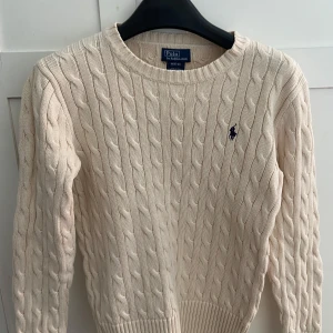 Beige kabelstickad tröja Polo Ralph Lauren - Kabelstickad beige tröja från Polo Ralph Lauren i 100% bomull. Klassisk rund halsringning, ribbade muddar och det ikoniska broderade logotypen på bröstet. Perfekt för dig som gillar en tidlös och clean stil. Storlek M för 10-12 åringar men funkar för vuxen storlek S/XS. Inga defekter 