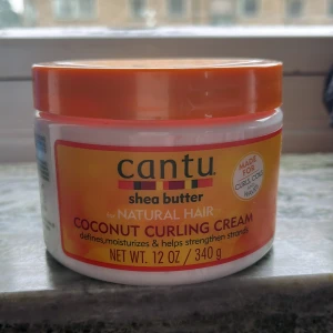 Cantu Coconut Curling Cream 340g - Cantu Coconut Curling Cream med sheasmör är en vit, krämig stylingprodukt för naturligt lockigt hår. Kommer i en rund plastburk med orange lock och etikett. Gammal förpackning 