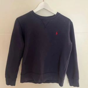 Mörkblå sweatshirt från Polo Ralph Lauren - Klassisk mörkblå sweatshirt från Polo Ralph Lauren med rund hals och röd broderad logga på bröstet. Tröjan har långa ärmar och är tillverkad i mjuk bomull, perfekt för en chill och stilren look.
