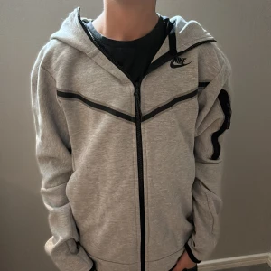En helt ny Nike tech fleece  - Snygg grå zip-hoodie från Nike med svarta detaljer och dragkedja hela vägen. Jackan har huva, lång ärm och coola svarta linjer över bröstet och på ärmen. Perfekt för chill dagar eller när du vill ha en sportig look.
