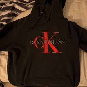Svart Calvin Klein hoodie med röd logga - Svart hoodie från Calvin Klein Jeans med stor röd CK-logga och text på bröstet. Tröjan har huva med snörning och en klassisk känguruficka framtill. Perfekt för dig som gillar streetstyle och vill sticka ut med en snygg logga.