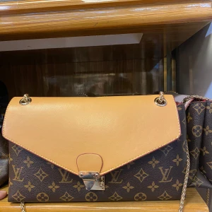  axelväska monogram - Snygg axelväska med klassiskt monogrammönster i brunt och beige. Väskan har guldfärgad kedjerem och lock i ljusbrunt skinn med metallspänne framtill. Perfekt för dig som vill ha en ikonisk och trendig accessoar.