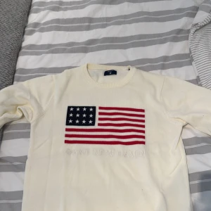 Vit stickad tröja från Gant - Vit stickad tröja från Gant med broderad amerikansk flagga på bröstet och texten 'GANT NEW HAVEN' under. Tröjan har rund halsringning och långa ärmar. Perfekt för dig som gillar klassisk och preppy stil.