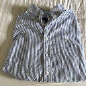 Randig skjorta från Gant - Snygg skjorta från Gant med blå och vita smala ränder, klassisk button down-krage och broderad logga på bröstet. Skjortan är långärmad och har vita knappar framtill. Perfekt för dig som gillar stilrena och tidlösa plagg. Bomull.